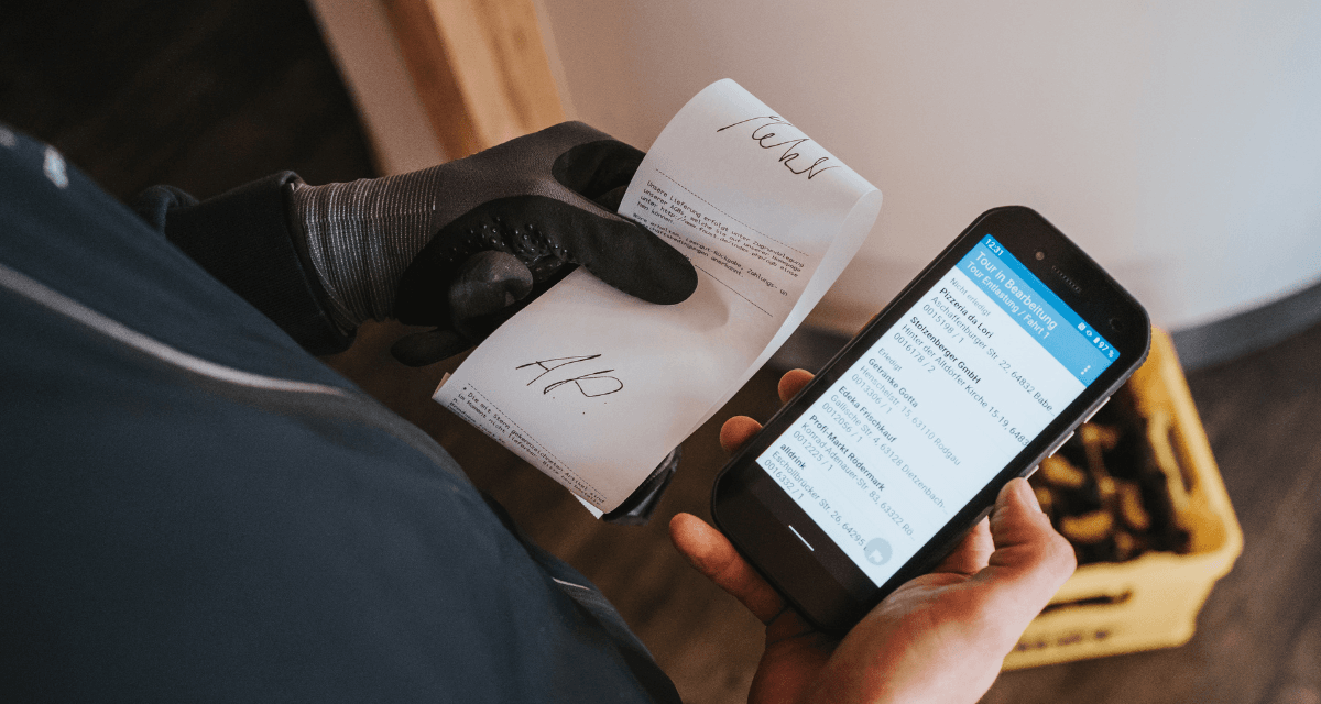 Ein Mann sitzt in einem Fahrzeug und bedient einen mobilen Drucker' w%E4hrend er einen frisch gedruckten Beleg entgegenzieht. Eine weitere Person steht daneben und h%E4lt das Papier fest.