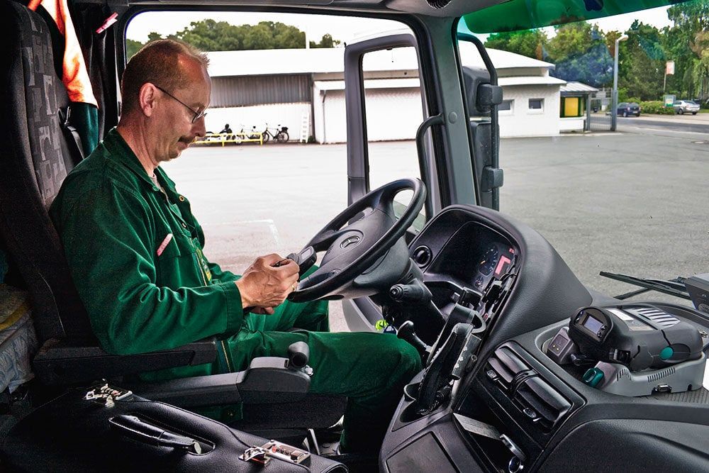 Ein%20Fahrer%20sitzt%20in%20der%20Kabine%20eines%20Lastwagens%20und%20bedient%20ein%20mobiles%20Ger%E4t%2C%20das%20auf%20dem%20Armaturenbrett%20liegt.%20Er%20tr%E4gt%20einen%20gr%FCnen%20Arbeitsanzug%20und%20konzentriert%20sich%20auf%20die%20Eingabe.