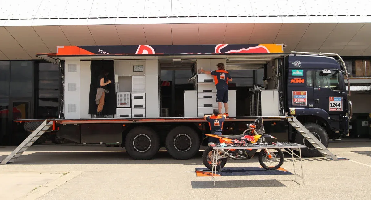 KTM racing team support truck with extended side panels revealing mobile workshop interior, crew members preparing equipment, and an orange KTM dirt bike displayed on a stand in front at a motorsport venue